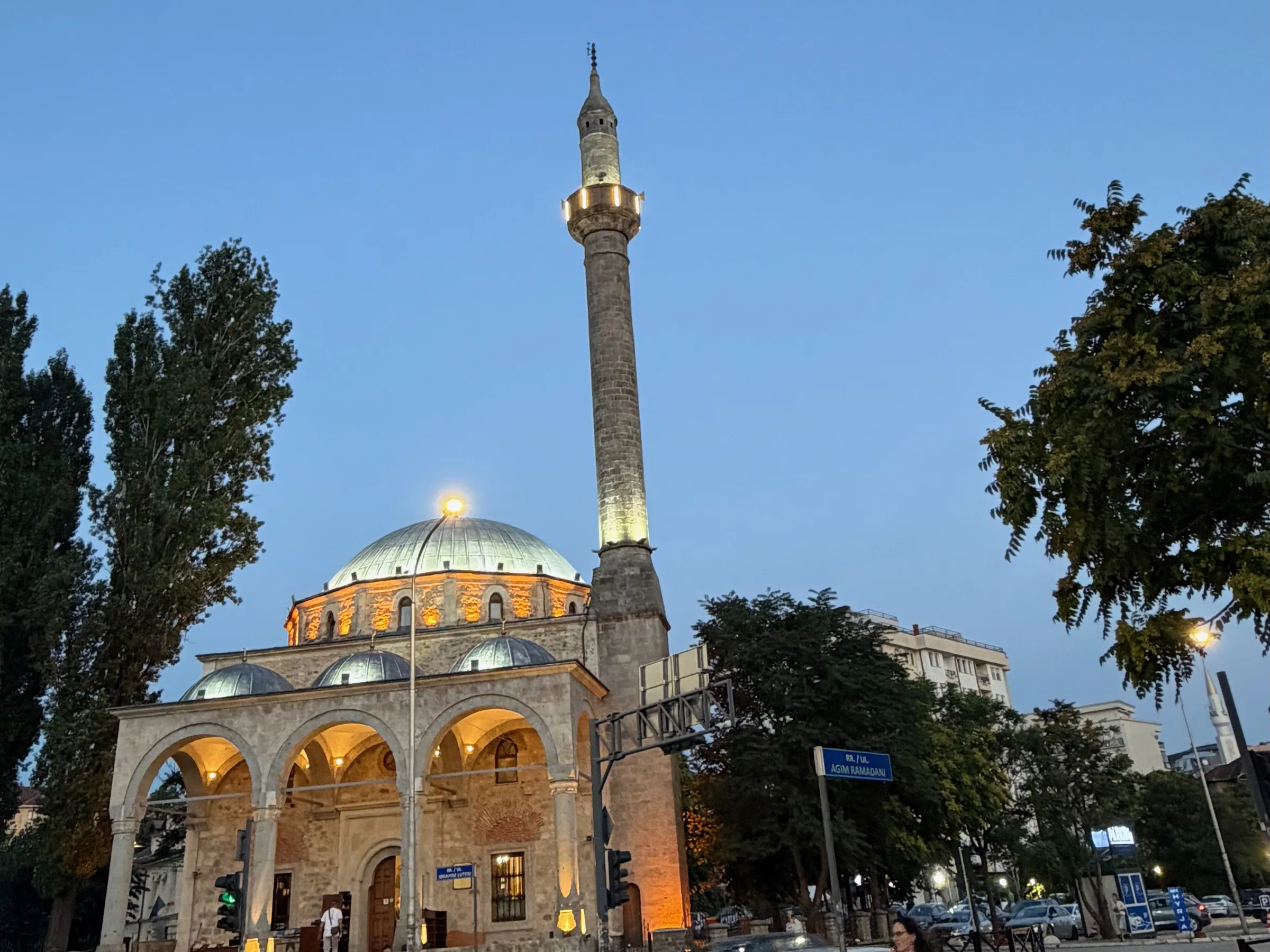 A domed Ottoman-era mosque in Pristina at dusk, its arcade and dome lit from below, minaret rising against a deep blue sky, with a street sign and evening traffic at the right of the frame.